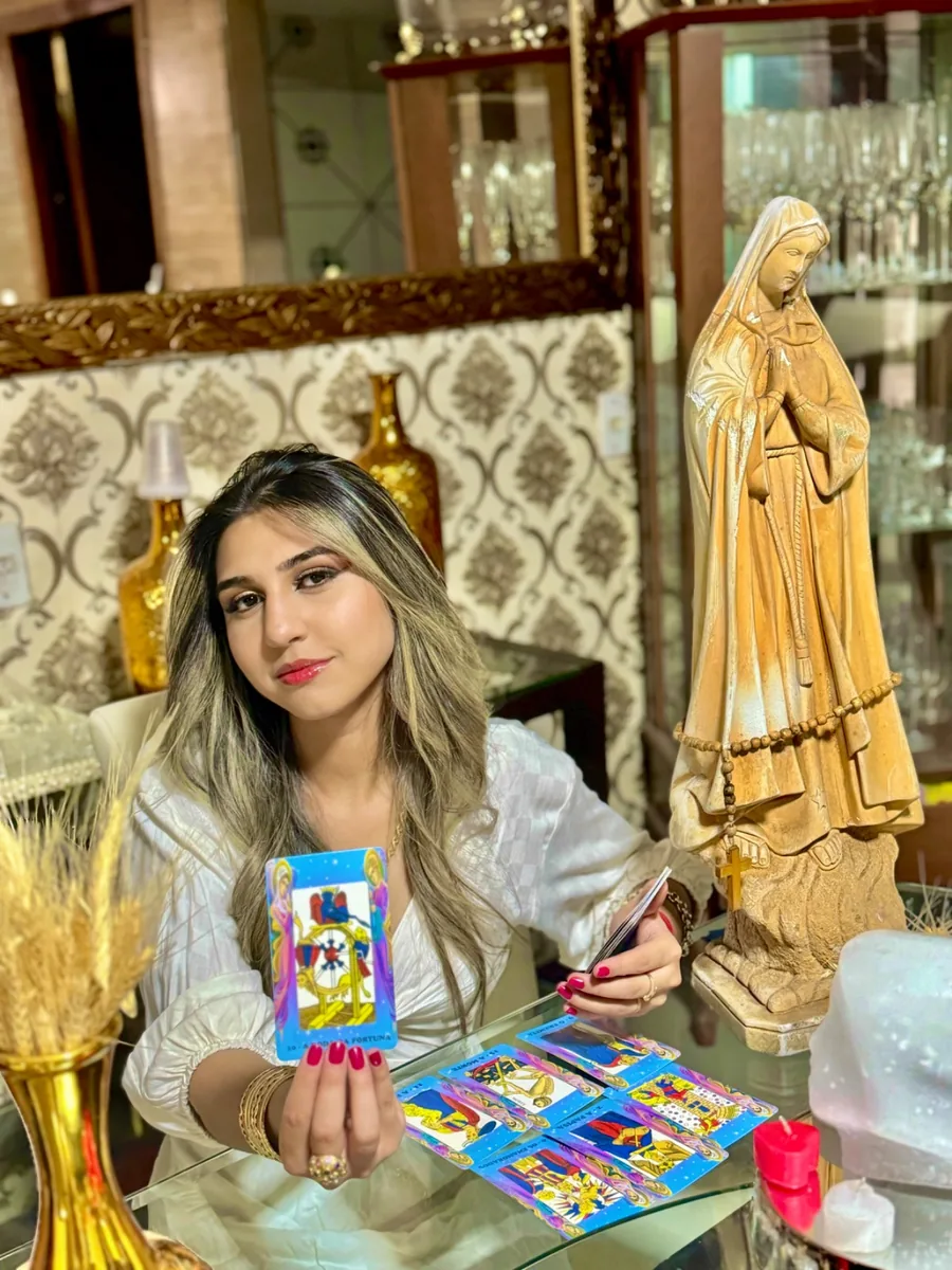 Amanda com blusa branca mostrando a carta A Roda da Fortuna, com outras cartas de tarô sobre a mesa.