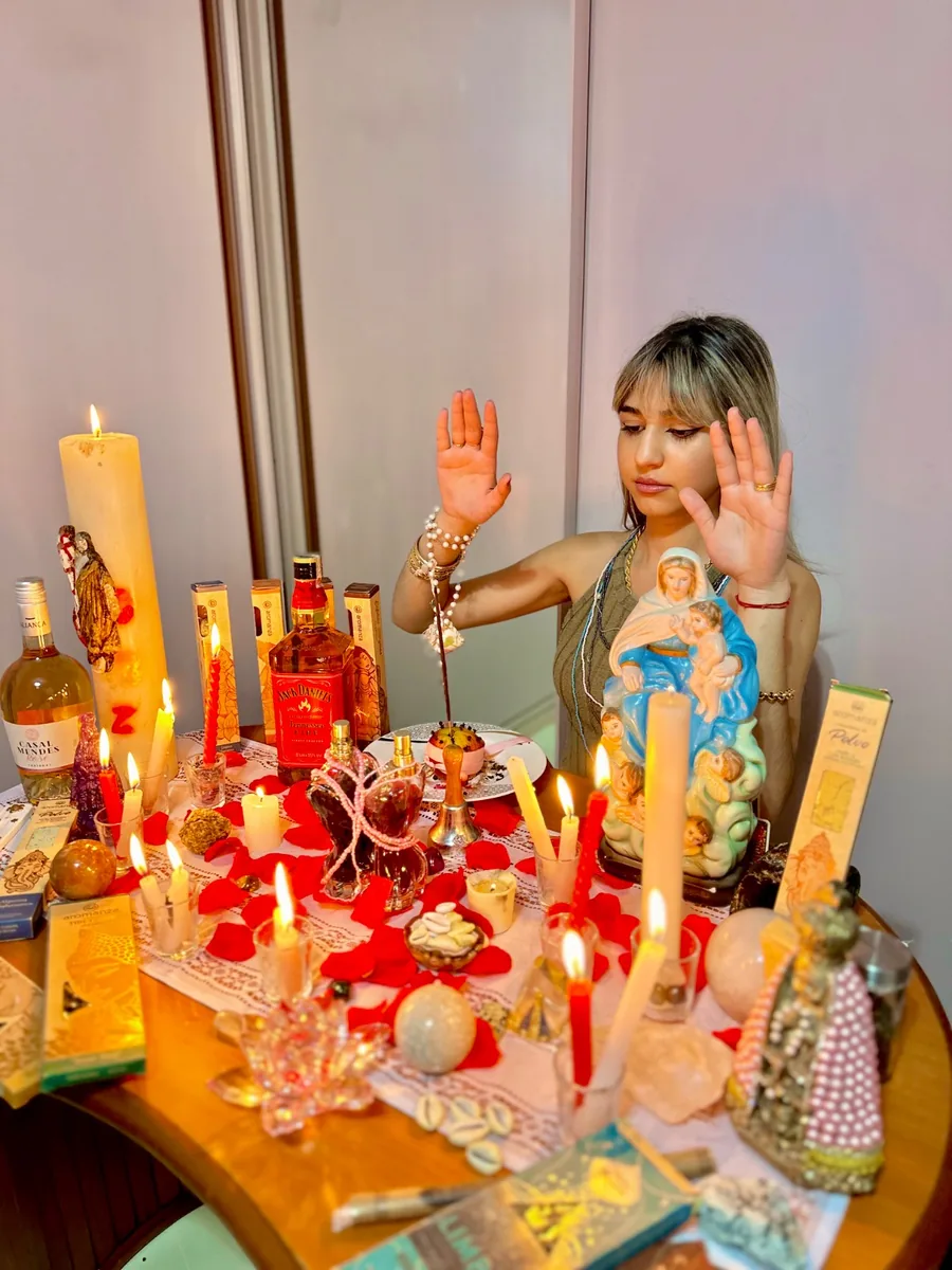 Amanda com as mãos erguidas diante de uma mesa com velas, pétalas, incensos e uma imagem de Nossa Senhora.